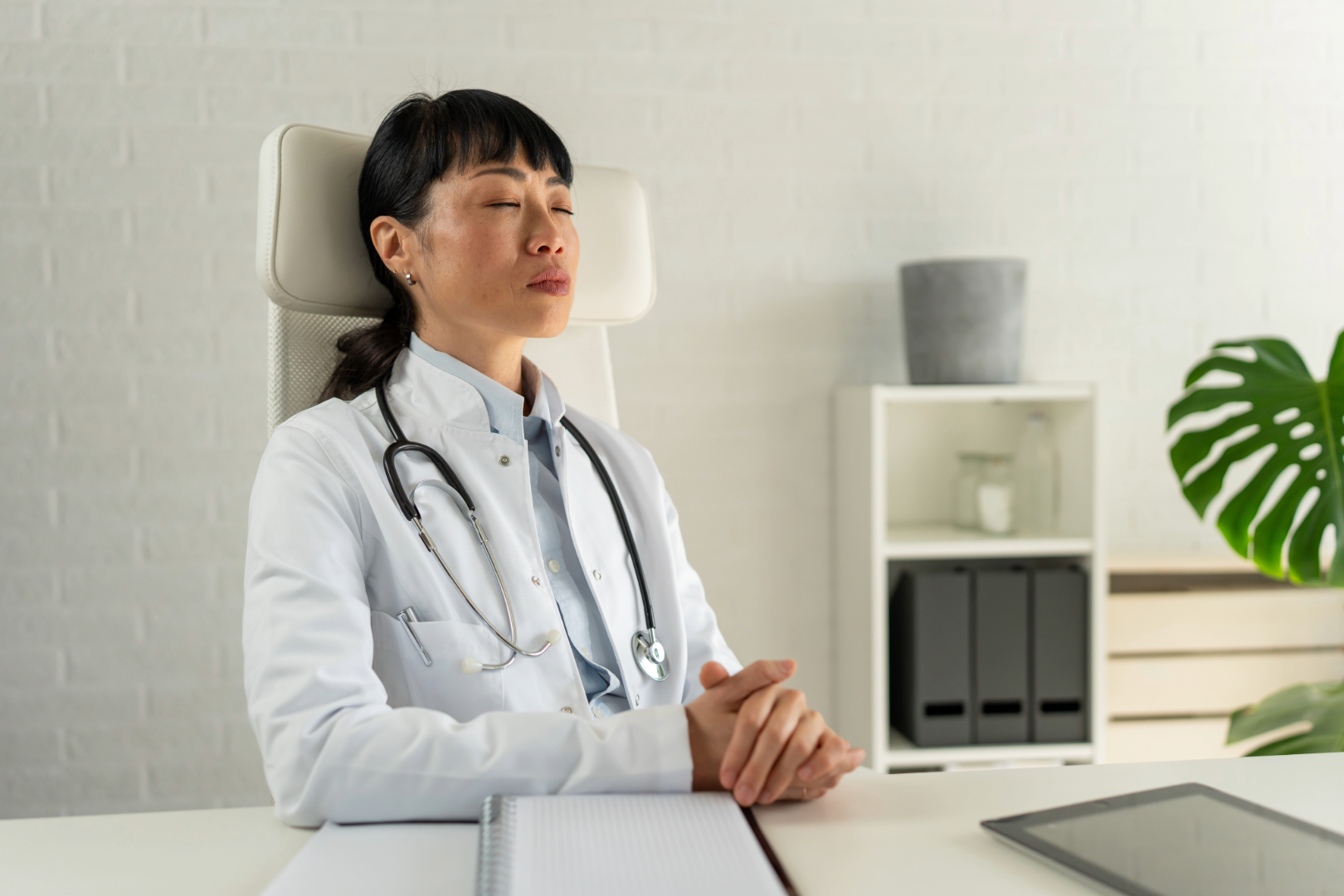 A clinician pausing with eyes closed for a quiet moment at her desk during the workday